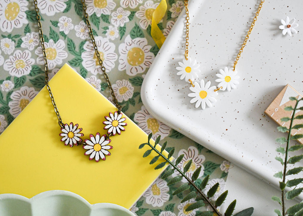 Two daisy chain necklaces displayed on a floral patterned surface and a white ceramic dish. One necklace has white acrylic daisies with a yellow painted center and gold plated chain, and the other has wooden daisies with white and pink details on a bronze chain.
