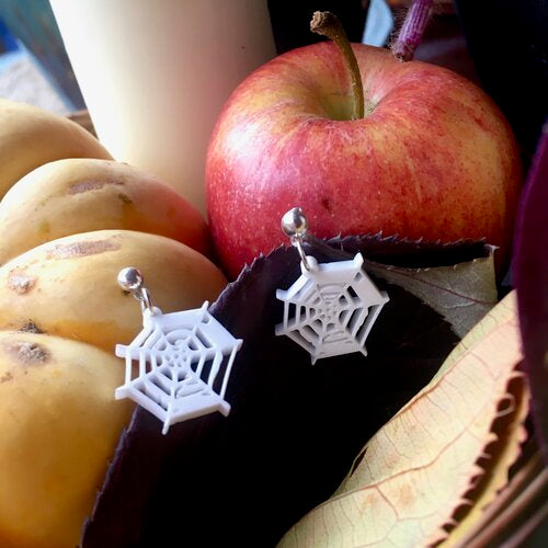 Laser cut white acrylic cobweb earrings displayed in front of an apple and leaves, suggesting an autumnal theme.
