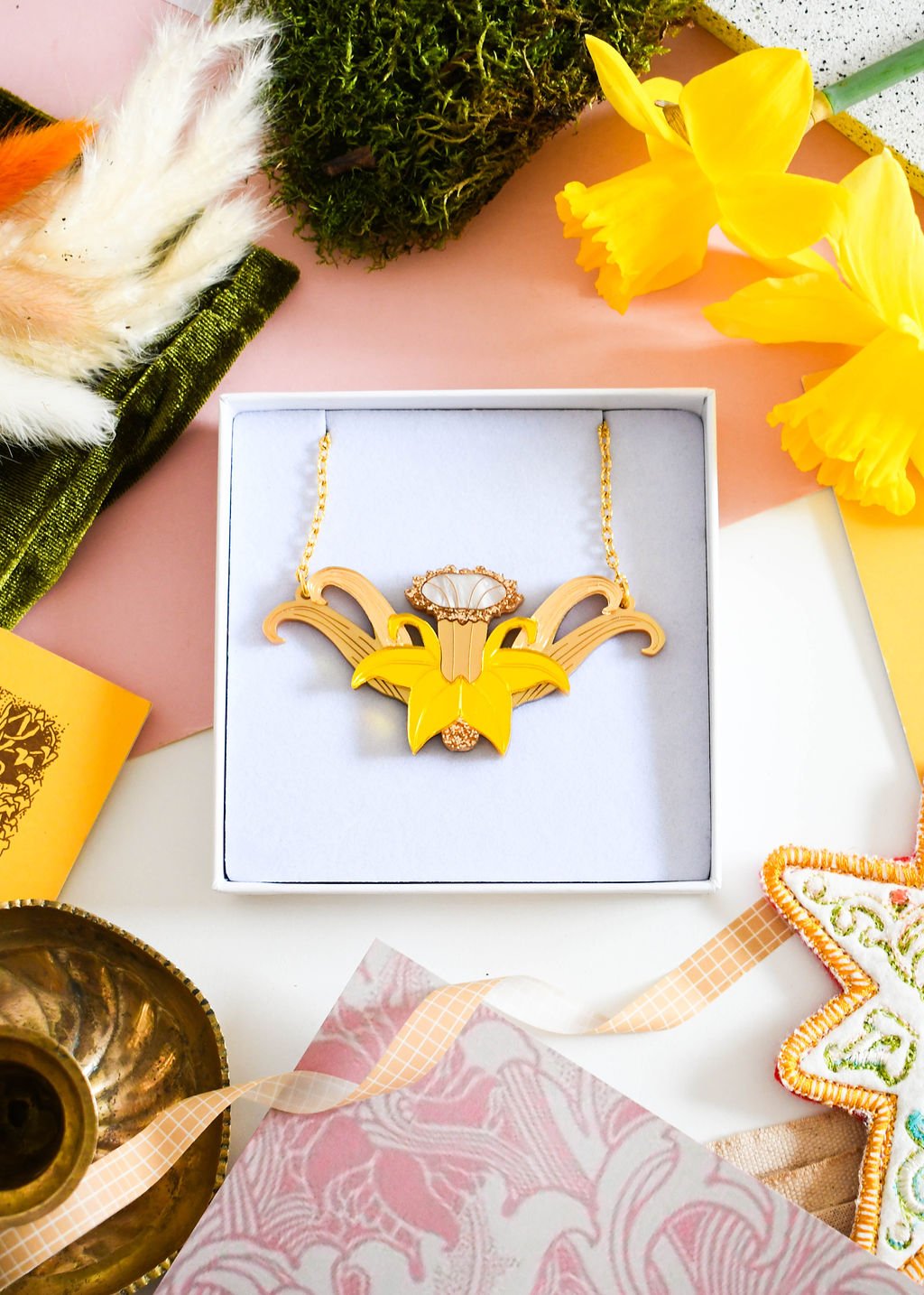 A necklace designed in an Art Nouveau style with yellow mirrored petals and gold-plated chain, displayed in a box with daffodil flowers and other prop decorations.