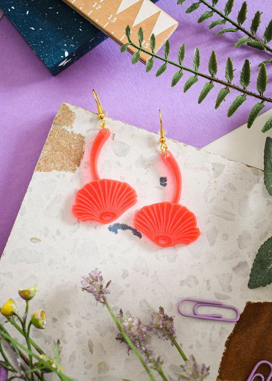 A pair of pink mushroom-shaped earrings with gold hooks, displayed on a piece of paper with foliage.
