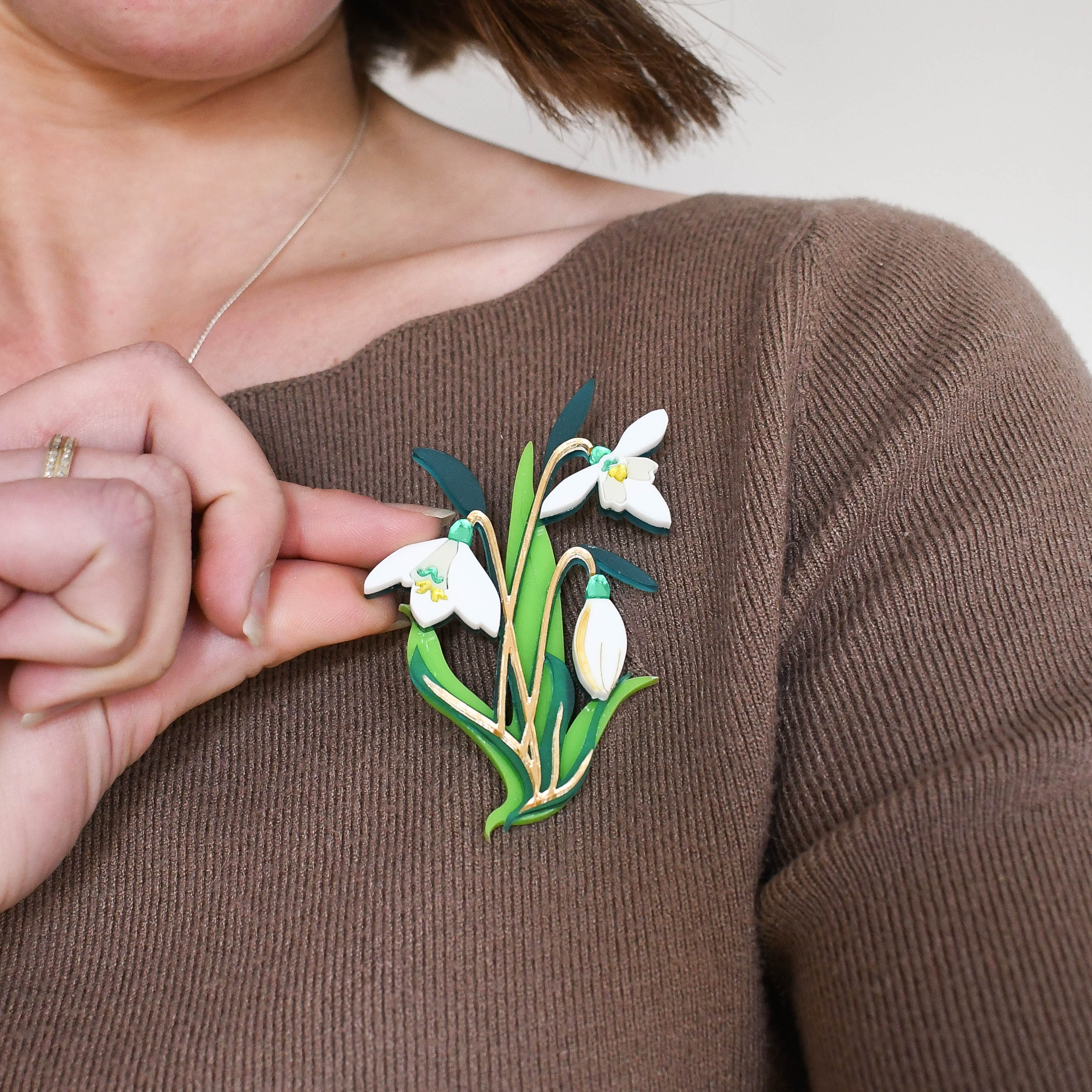 A person wearing a brown sweater with a brooch depicting snowdrop flowers in green, white, and yellow colors pinned to the lapel.