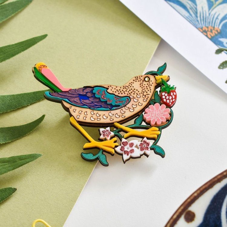 A colorful brooch depicting a bird with a strawberry in its beak, surrounded by floral patterns, placed on a green fabric background.