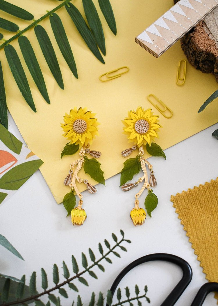 A pair of vibrant yellow sunflower statement earrings displayed on a table with various objects, including a pair of scissors, paper clips, and a craft knife.
