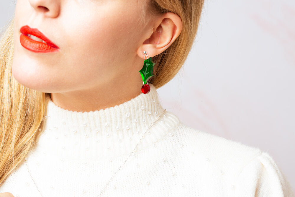 A person wearing a white turtleneck sweater and red lipstick, showcasing dangling earrings with green and red acrylic berries.