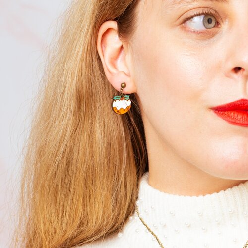 A person wearing Christmas pudding-shaped earrings, with the earrings visible against their ear. The earrings are colorful and designed to resemble a Christmas pudding cake.